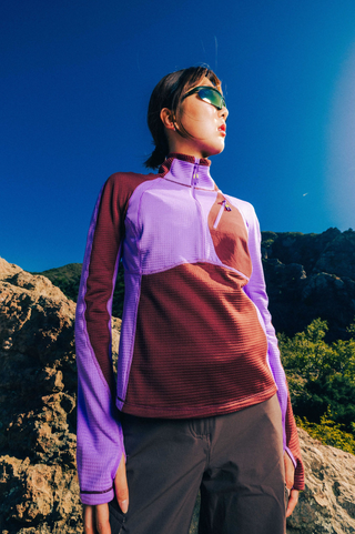 Woman wearing a SENIQ Joytrek Midlayer Fleece in Brambleberry standing on a mountain with a clear blue sky.