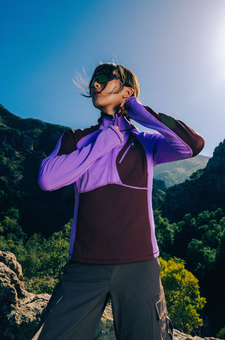 Woman wearing a SENIQ Joytrek Midlayer Fleece in Brambleberry standing on a mountain with a clear blue sky.