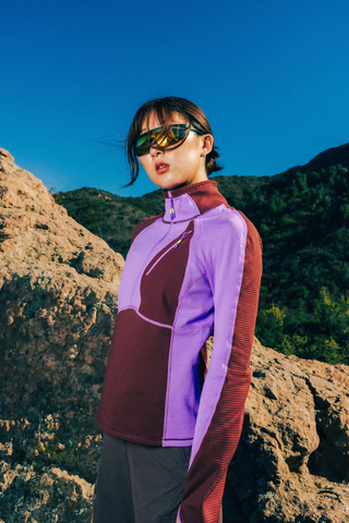 Woman wearing a SENIQ Joytrek Midlayer Fleece in Brambleberry standing on a mountain with a clear blue sky.