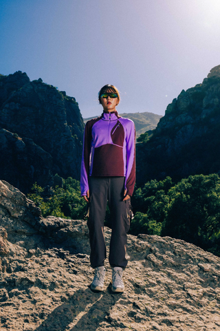 Woman wearing a SENIQ Joytrek Midlayer Fleece in Brambleberry standing on a mountain with a clear blue sky.